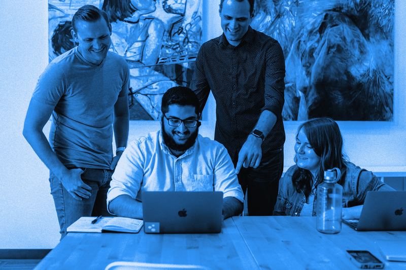 Four coworkers smiling around a laptop at a shared table